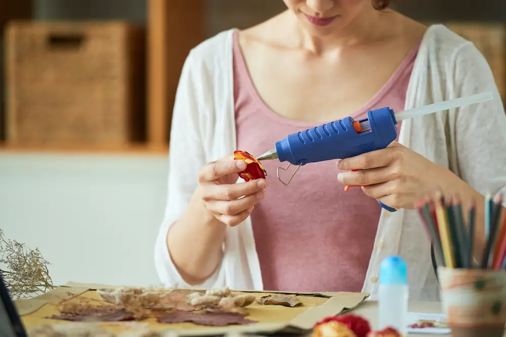 Close-up shot of a woman using hot glue gun while wrapped up in making floral decor, interior of living room on background/Getty Images
